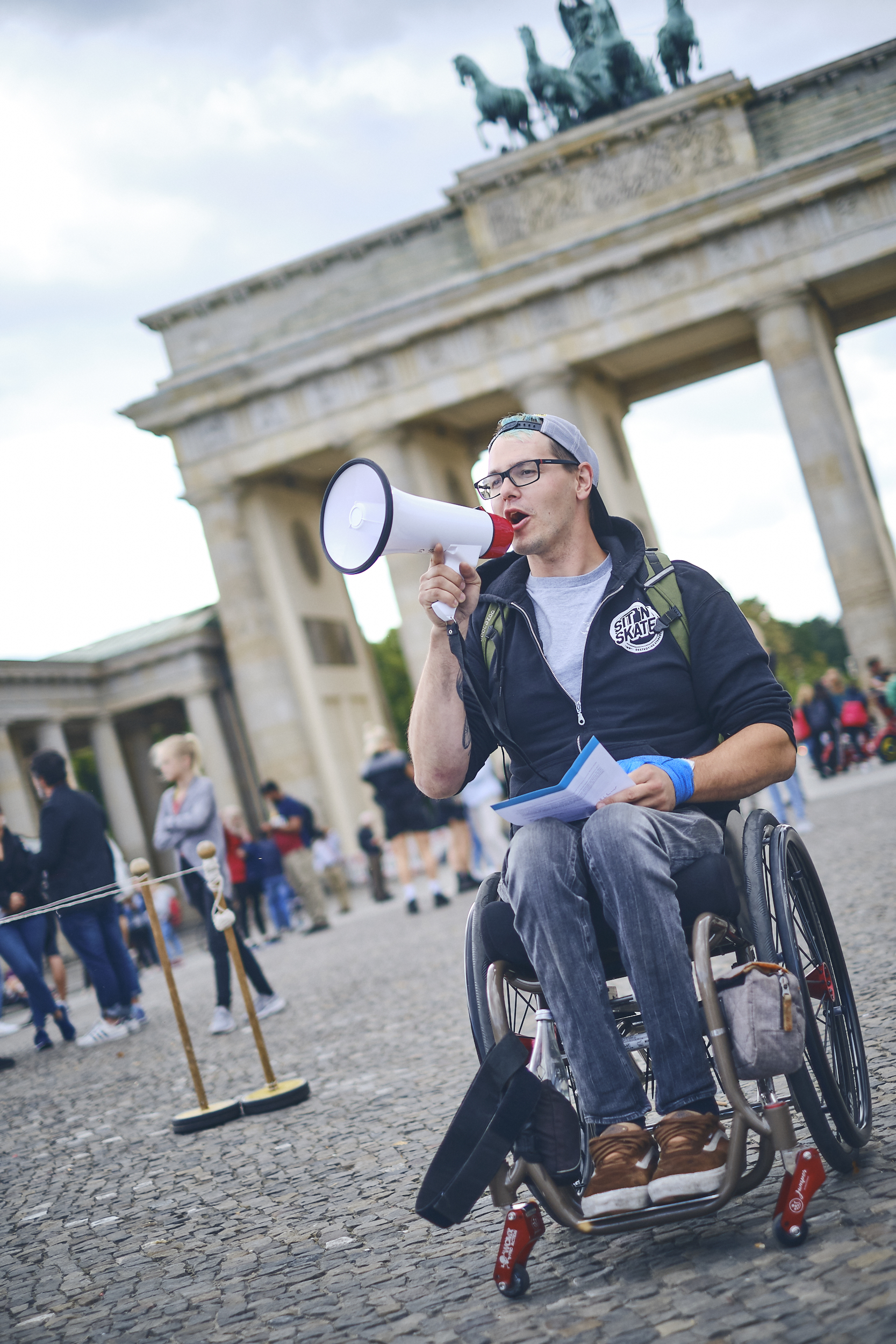 ein weißer Rollstuhlfahrer steht mit Megaphon vor dem Brandenburger Tor