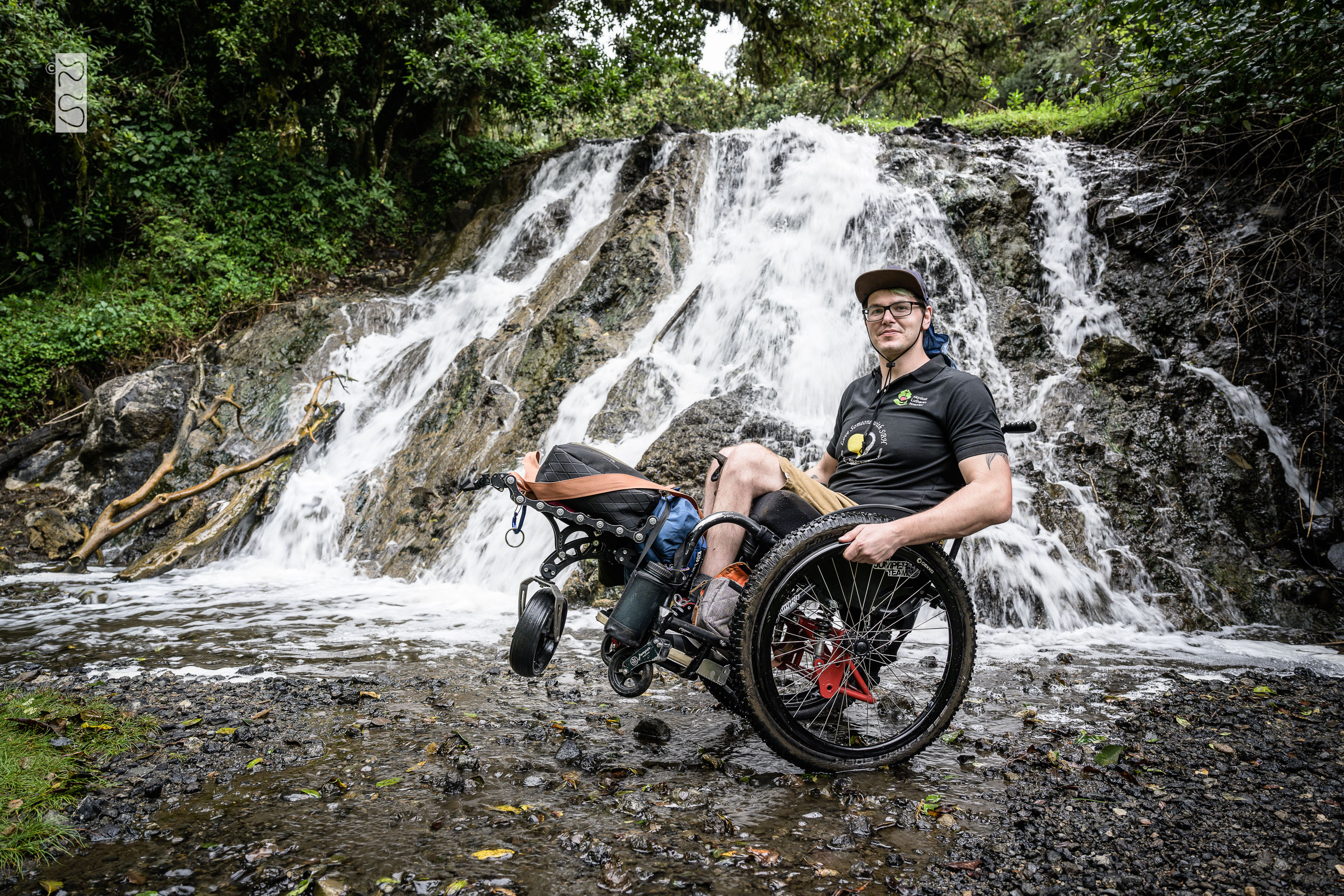 David posiert mit seinem Rollstuhl vor einem Wasserfall