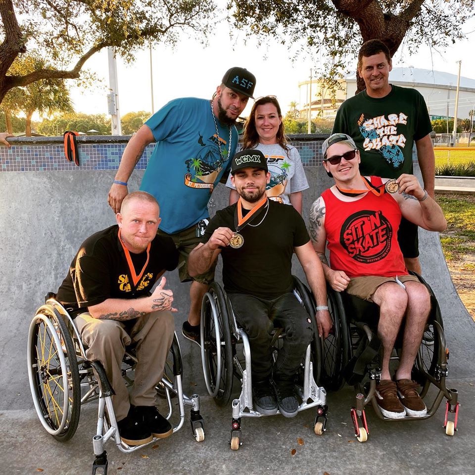Auf dem Bild zeigen drei Rollstuhlfahrer ihre Medaillen hoch, dahinter stehen zwei Männer und eine Frau vor einer Beton Skate Rampe