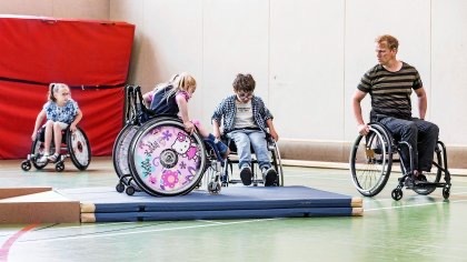 Drei Kinder und ein Erwachsener in Rollstühlen in einer Turnhalle. Zwei Kinder trainieren an und auf Matten