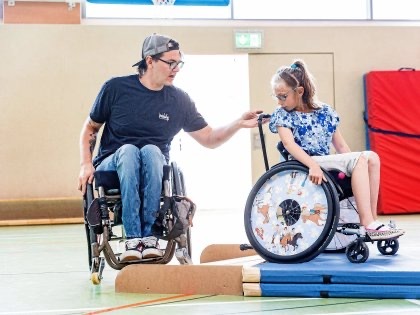 Ein Mann und ein Mädchen, beide in Rollstühlen, in einer Turnhalle. Das Mädchen steht auf Matten, der Mann sichert sie von hinten ab.