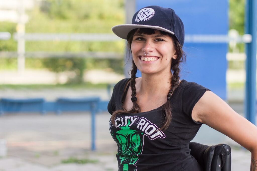 Ein Foto von Lisa aus dem Jahr 2014. Sie trägt ein Cap, Zöpfe und ein Top wo die Band Rat City Riot drauf steht.