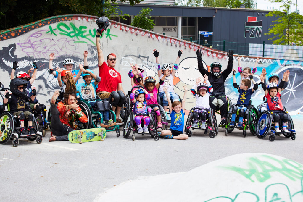 Gruppenfoto mit vielen Rollstuhlfahrern, davon viele Kinder, mit Helmen in einem Skatepark. 
