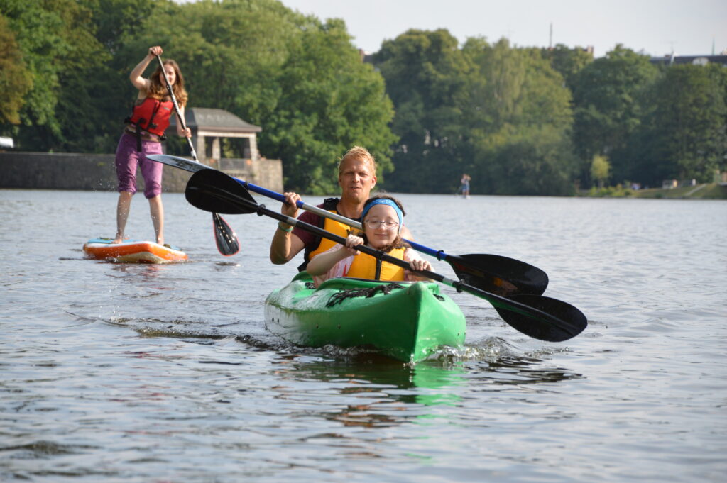 Drei Personen paddeln: eine Frau stehend auf einem Stand-Up Paddeling Board und ein Mann und ein Mädchen in einem Kanu