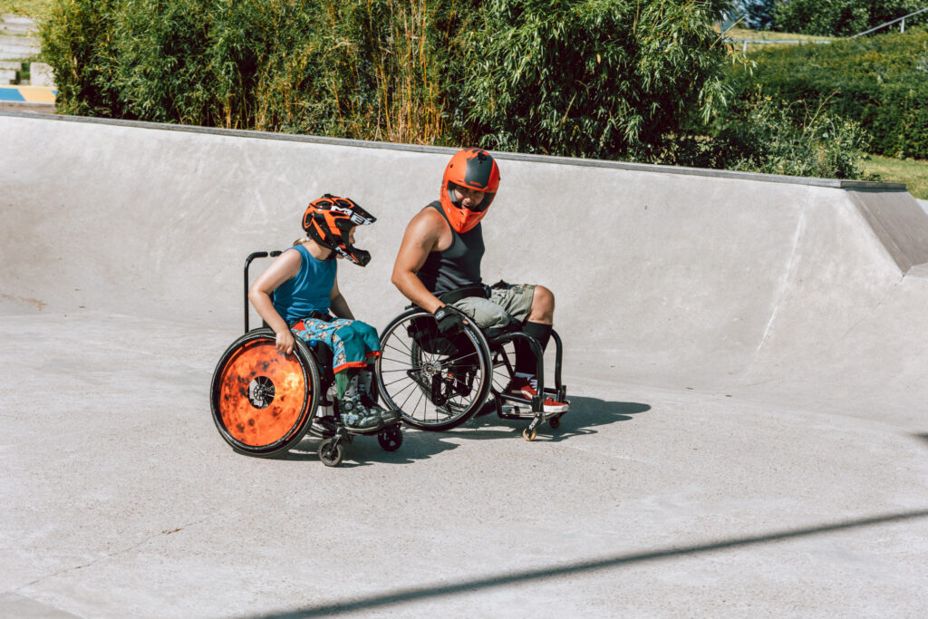 Ein Junge und ein Mann, bei im Rollstuhl, mit Helmen in einem Skatepark