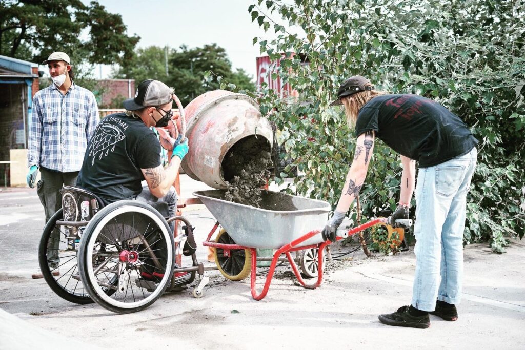 Ein Mann hält eine Schubkarre während ein anderer Mann im Rollstuhl Beton aus einem Betonmischer in die Schubkarre gießt