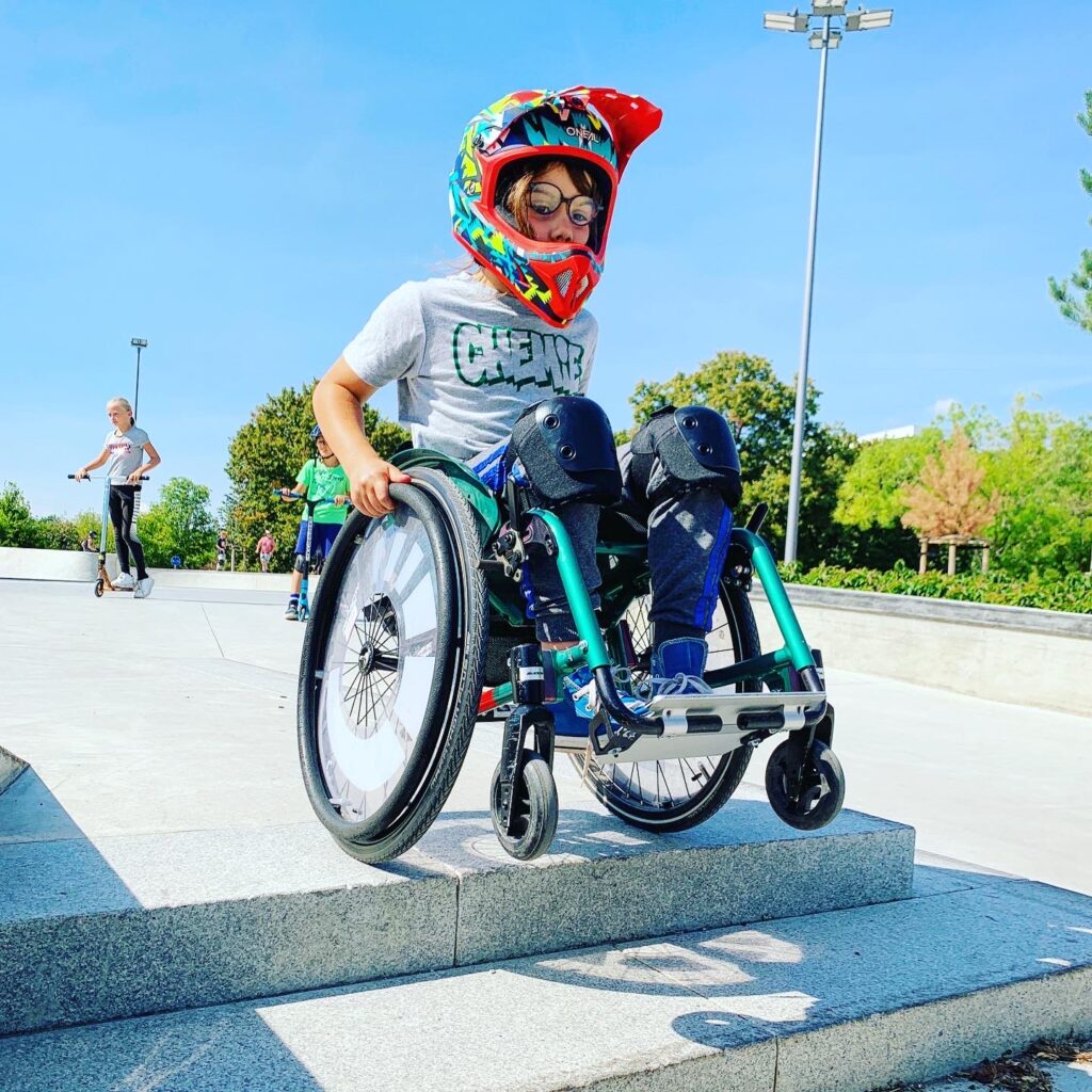 ein Junge im Rollstuhl mit Fullface Helm balanciert vor einer Treppe auf den Hinterrädern. 