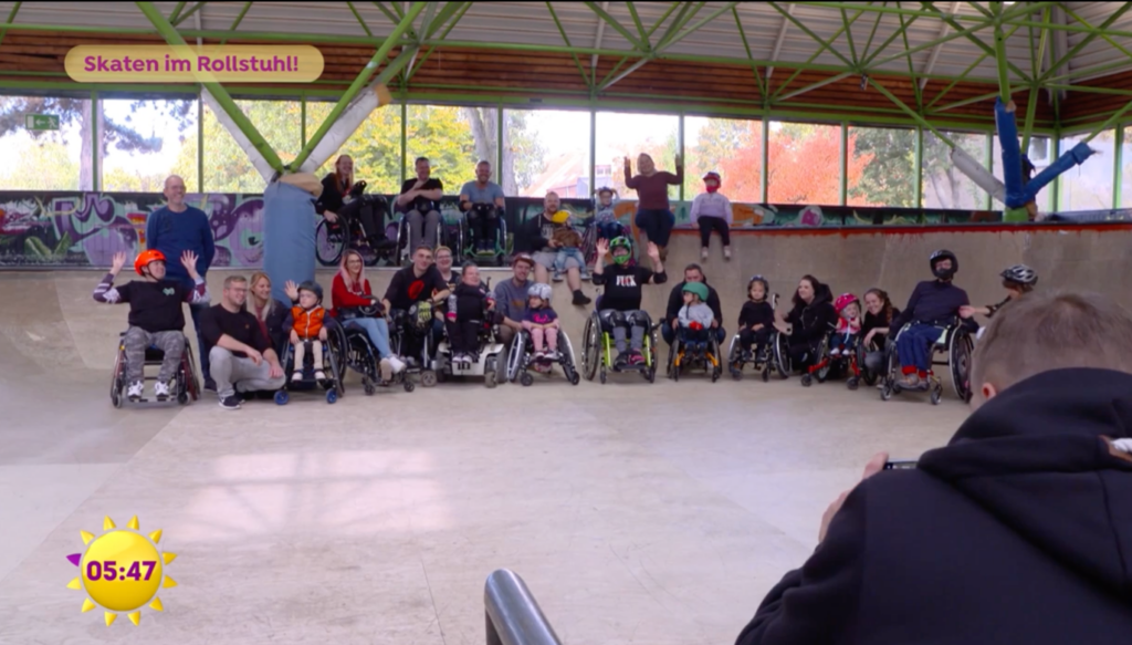 Screenshot vom Sat 1 Frühstücksfernsehen zum Thema "Skaten im Rollstuhl". Es wird ein Gruppenbild in der Skatehalle Dortmund mit vielen Rollstuhlfahrern gezeigt. Davon viele sind viele Kinder