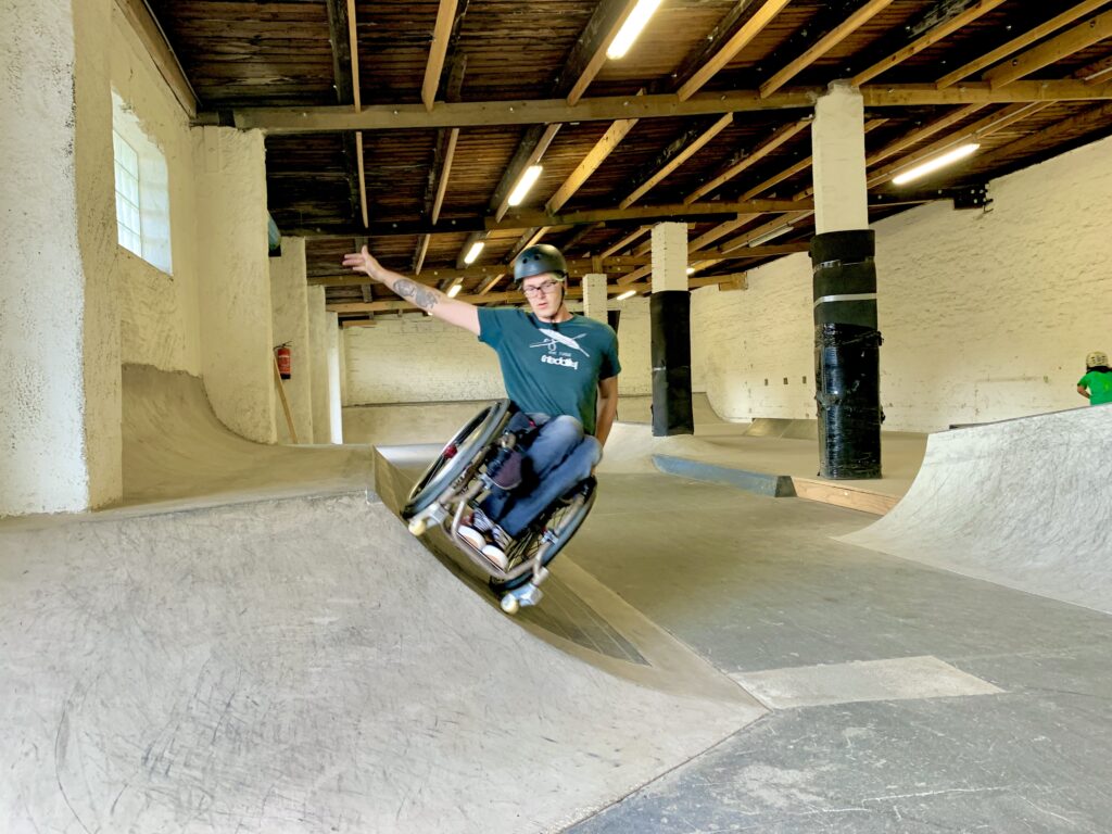 Ein Rollstuhlfahrer fährt in einer Skatehalle an einer Beton rampe seitlich entlang und streckt den Arm aus um Balance zu halten. 