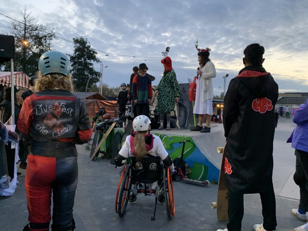 Verkleidete Person, davon ein Mädchen im Rollstuhl, in einem Skatepark. Einige stehen auf einer Rampe wie auf einer Bühne