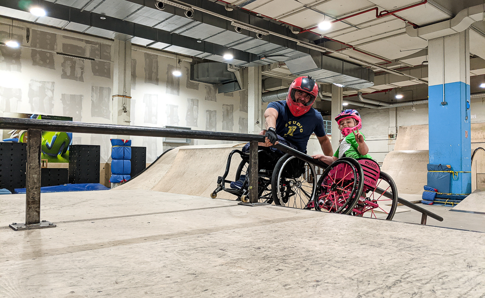 Ein erwachsener Rollstuhlfahrer und ein Mädchen im Rollstuhl in einer Skatehalle. Sie halten sich auf einer Schrägen an einem Geländer fest.