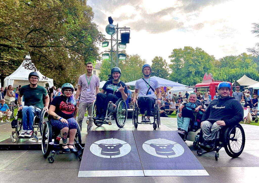 Eine Gruppe Rollstuhlfahrerinnen und Rollstuhlfahrer, sowie ein Fußgänger stehen auf einem mobilen Holzskatepark. Auf den Rampen sind Gorilla Gesichter als Logo und im Hintergrund Zelte und Zuschauer.