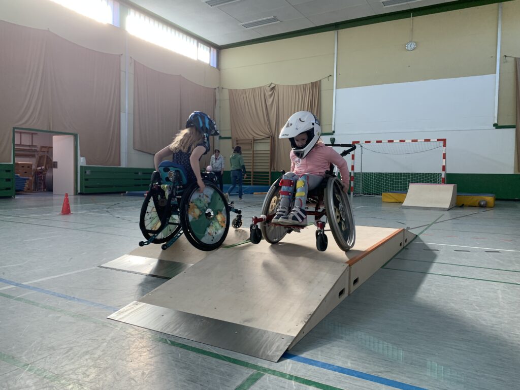 Zwei Mädchen im Rollstuhl in einer Turnhallte auf Skaterampen