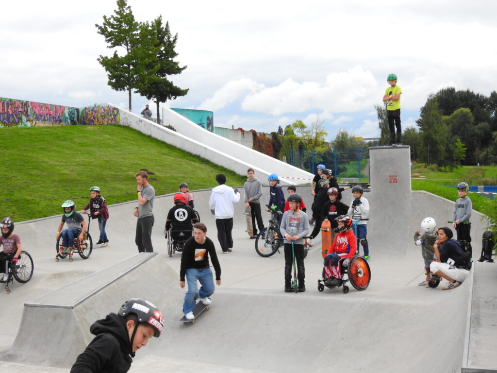 Eine kunterbunte Mischung im Skateboar, Skateboarder, Scooter und Rollstuhlfahrer verschiedenen Alters.
