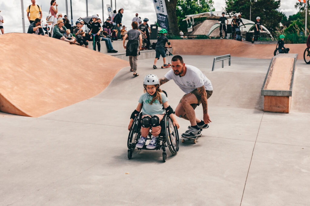 Ein Skateboarder fährt mit einem Mädchen im Rollstuhl durch den Skatepark.