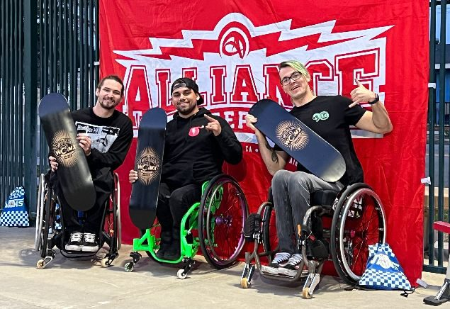 Drei Rollstuhlfahrer posieren vor einem roten Banner. 
