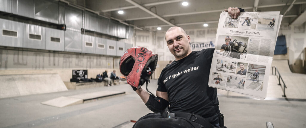 Björn steht in einer Skatehalle. In einer Hand hält er seinen Helm in der anderen eine Zeitung mit Fotos von ihm drin.