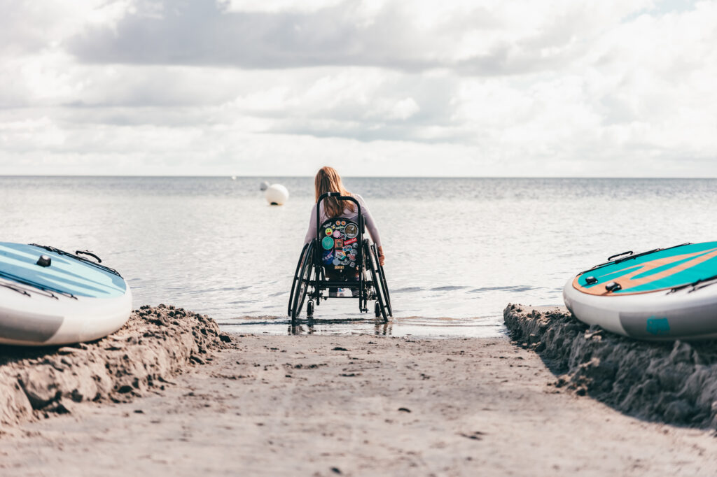 Ein Mädchen im Rollstuhl steht auf einem geräumten Weg am Strand, mit den Rädern im Meer. Links und rechts sind SUP Boards zu sehen.