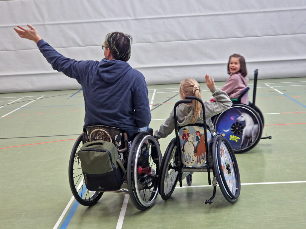 Zwei Kinder im Rollstuhl spielen Fange mit einem erwachsenen Rollstuhlfahrer in einer Sporthalle