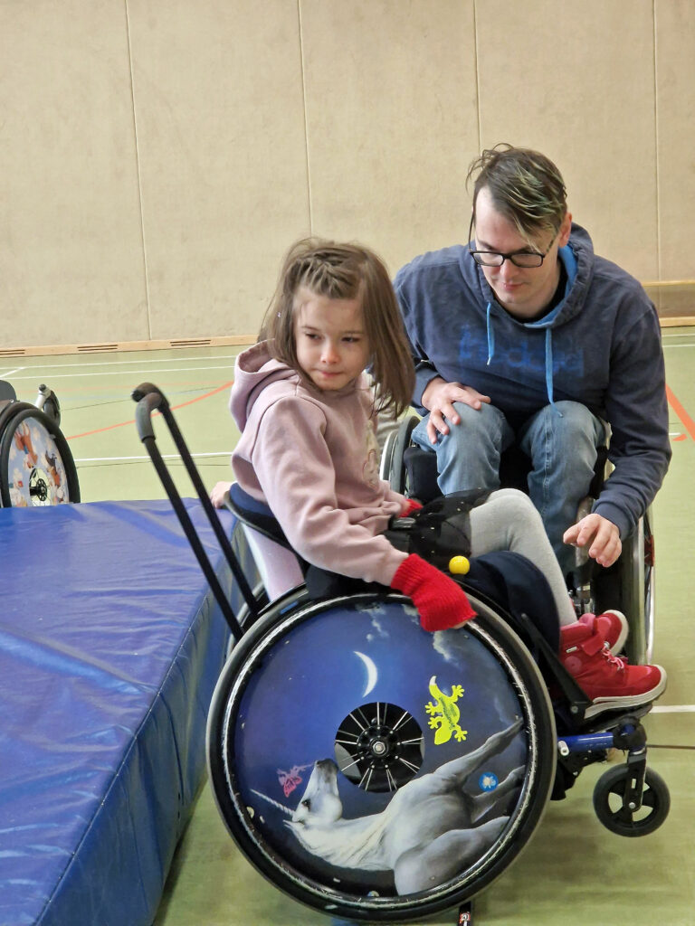 ein Mädchen im Rollstuhl übt auf den hinterrädern zu stehen. Hinter ihr ist eine Matte. Neben ihr ein erwachsener Rollstuhlfahrer.