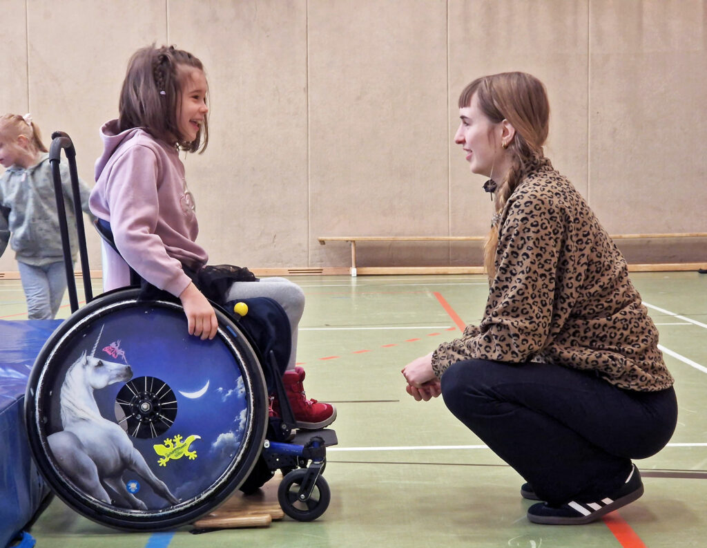 Eine Frau kniet vor einem Mädchen im Rollstuhl in der Sporthalle.