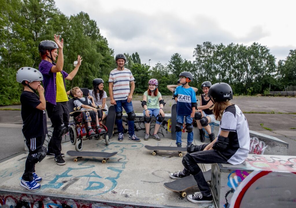 In einem Skatepark zeigt eine Frau Kindern mit und ohne Behinderung etwas mit den Händen. auf dem Boden stehen Skateboards. Ein Kind sitzt im Rollstuhl.