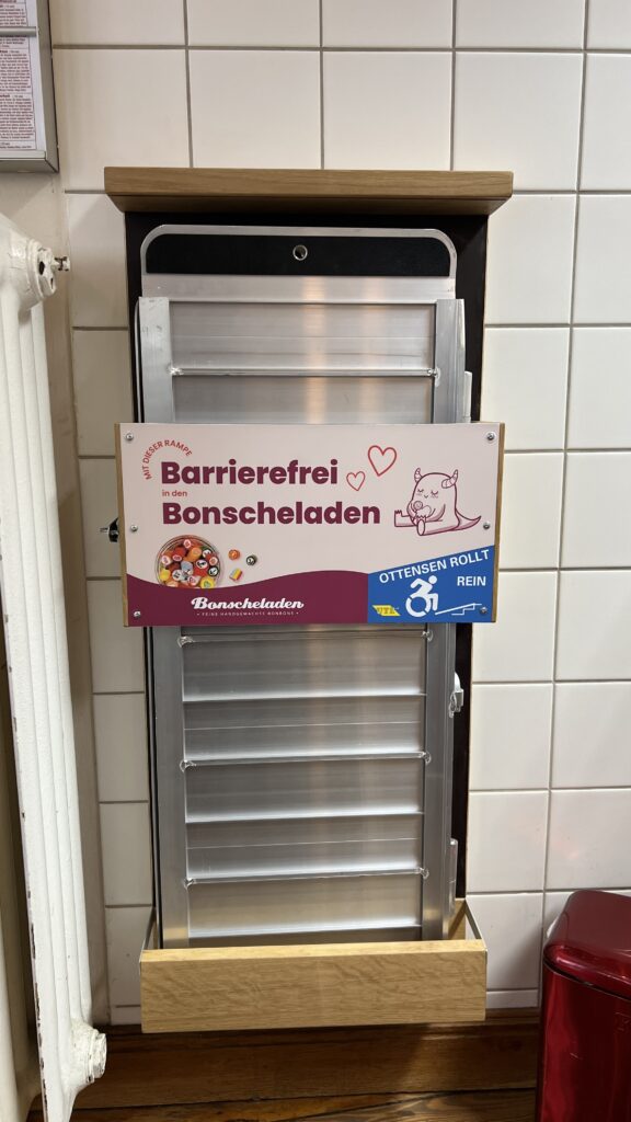 Eine Rollstuhlrampe in einer Holzhalterung mit einem Schild: Bonscheladen barrierefrei.