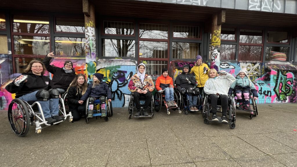 Eine Gruppe von Kindern, Jugendlichen und Erwachsenen mit und ohne Rollstuhl vor einer Graffiti Wand.