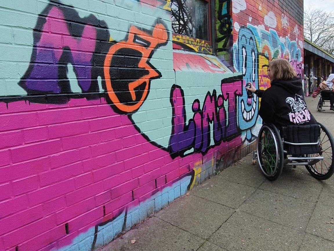 Man sieht eine Wand mit Graffiti, das Motiv ist No Limit, wobei das O ein Rollstuhl Symbol ist. Eine Frau im Rollstuhl arbeitet daran.
