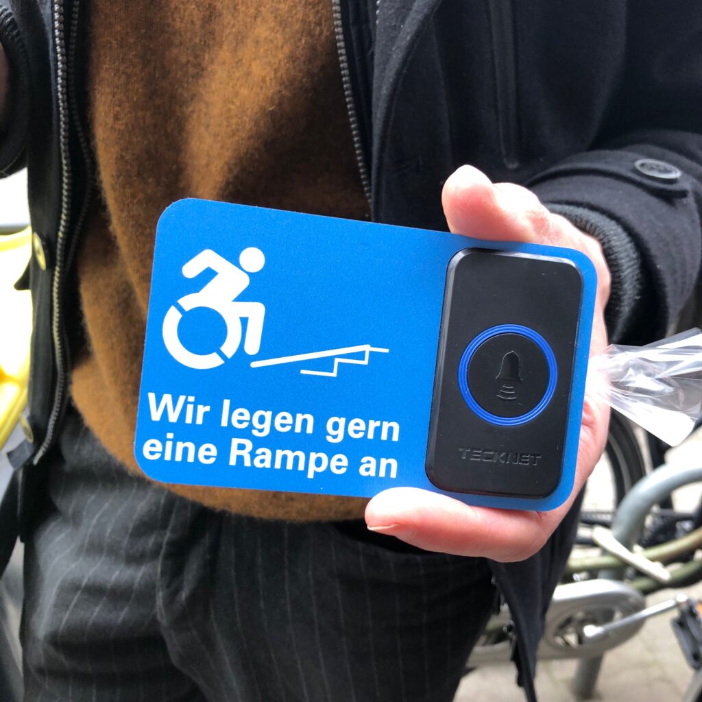 Jemand hält einen Klingelknopf in die Kamera mit einem Schild: An einer Schaufensterscheibe klebt ein Sticker mit einem Rollstuhlsymbol und einer angedeuteten Rampe über einer Treppe. Darunter steht "Wir legen eine Rampe an"