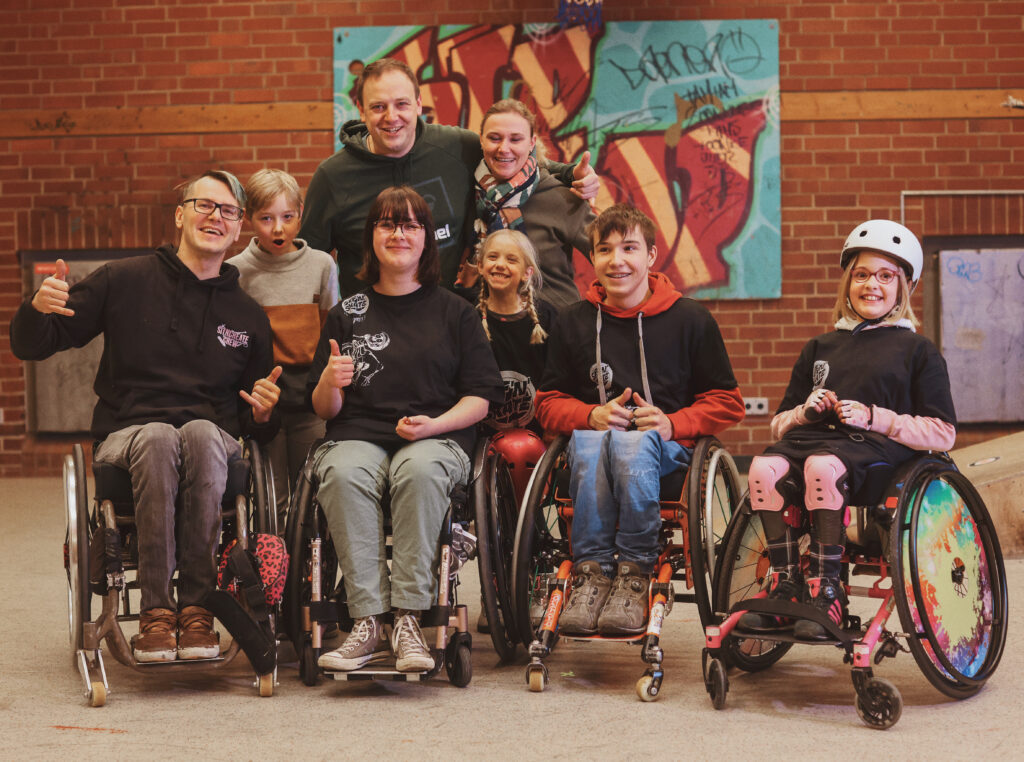 Eine Gruppe von Menschen stehen zusammen für ein Foto, ein Mädchen im Rollstuhl, Jugendliche im Rollstuhl, sowie ein Erwachsener im Rollstuhl und weitere zwei Erwachsene und zwei Kinder die Stehen. 