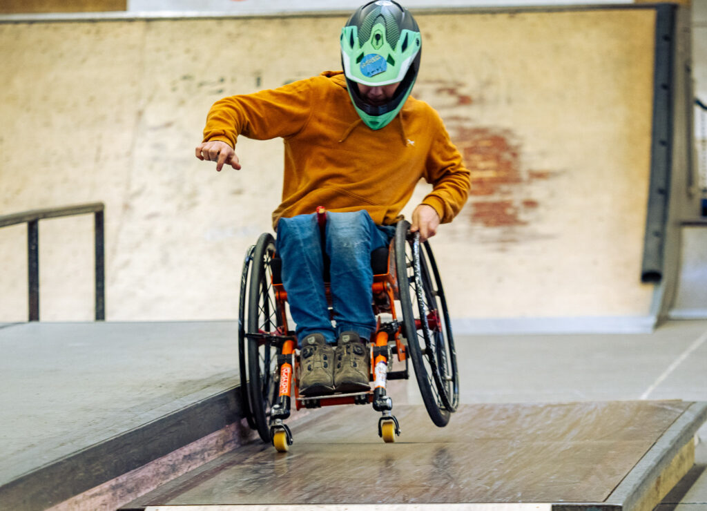  Ein Jugendlicher im Rollstuhl fährt im Skatepark auf einem Rad an eine Metallkante gelehnt.