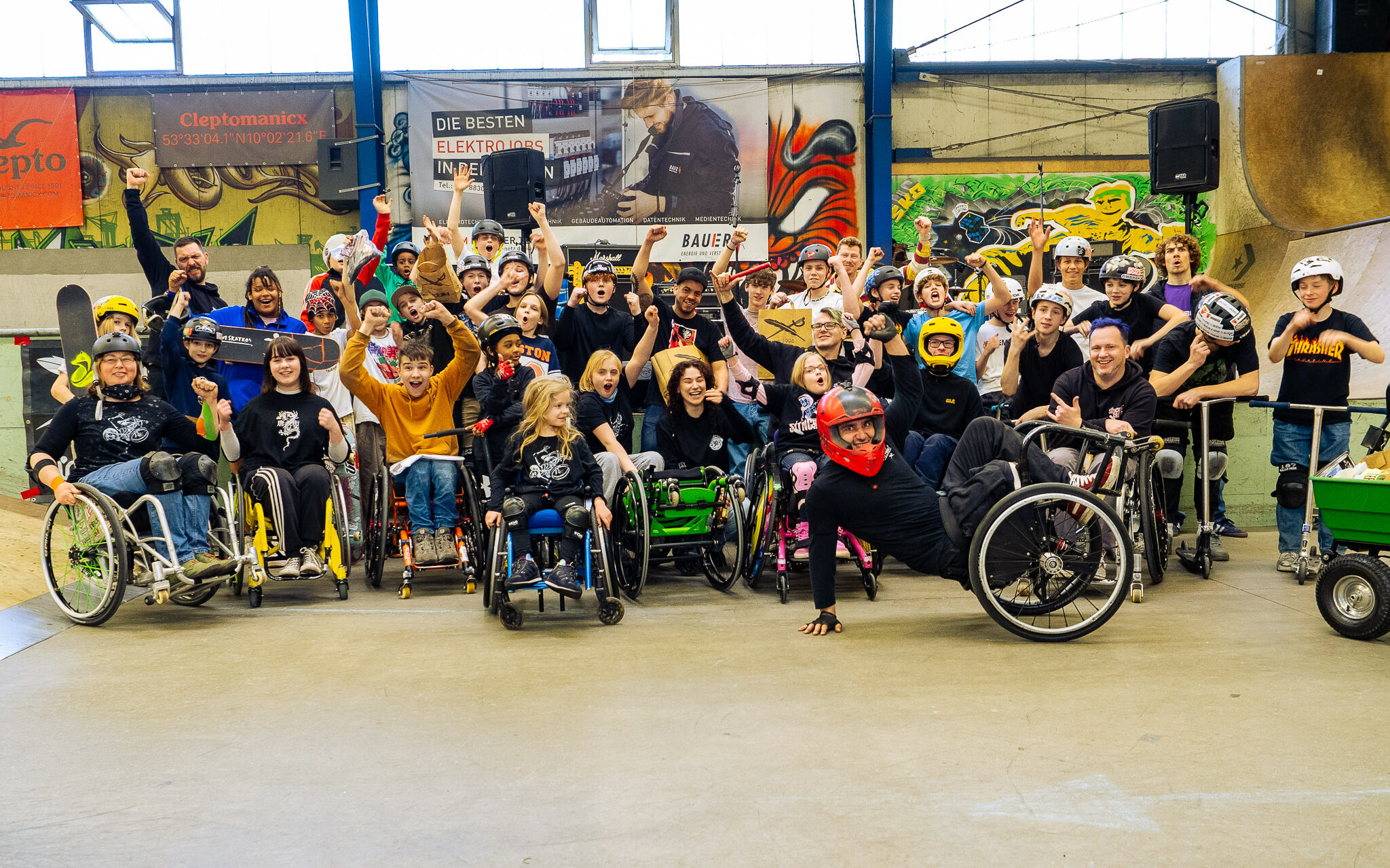 Gruppenbild in einer Skatehalle mit vielen Kindern, Jugendlichen und Erwachsenen in Rollstühlen, Scootern und Skateboards.