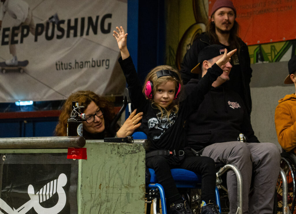 Ein Kind im Rollstuhl mit Ohrenschützer streckt die Arme nach oben zum jubeln.
