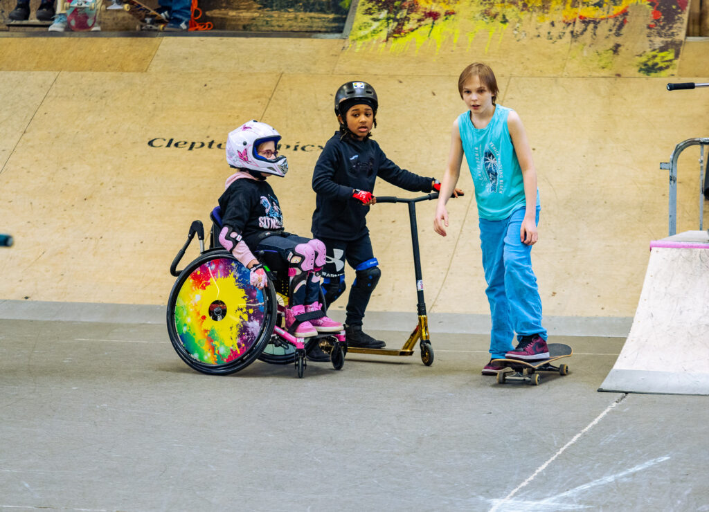 Drei Kinder in einer Skatehalle, ein Mädchen im Rollstuhl, ein Junge mit Scooter, ein Junge mit Skateboard