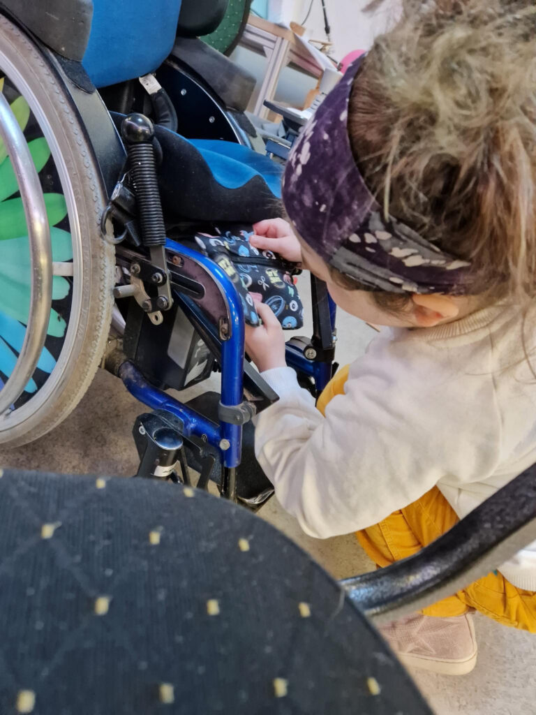 Ein Mädchen macht die fertige Tasche an ihrem Rollstuhl fest.