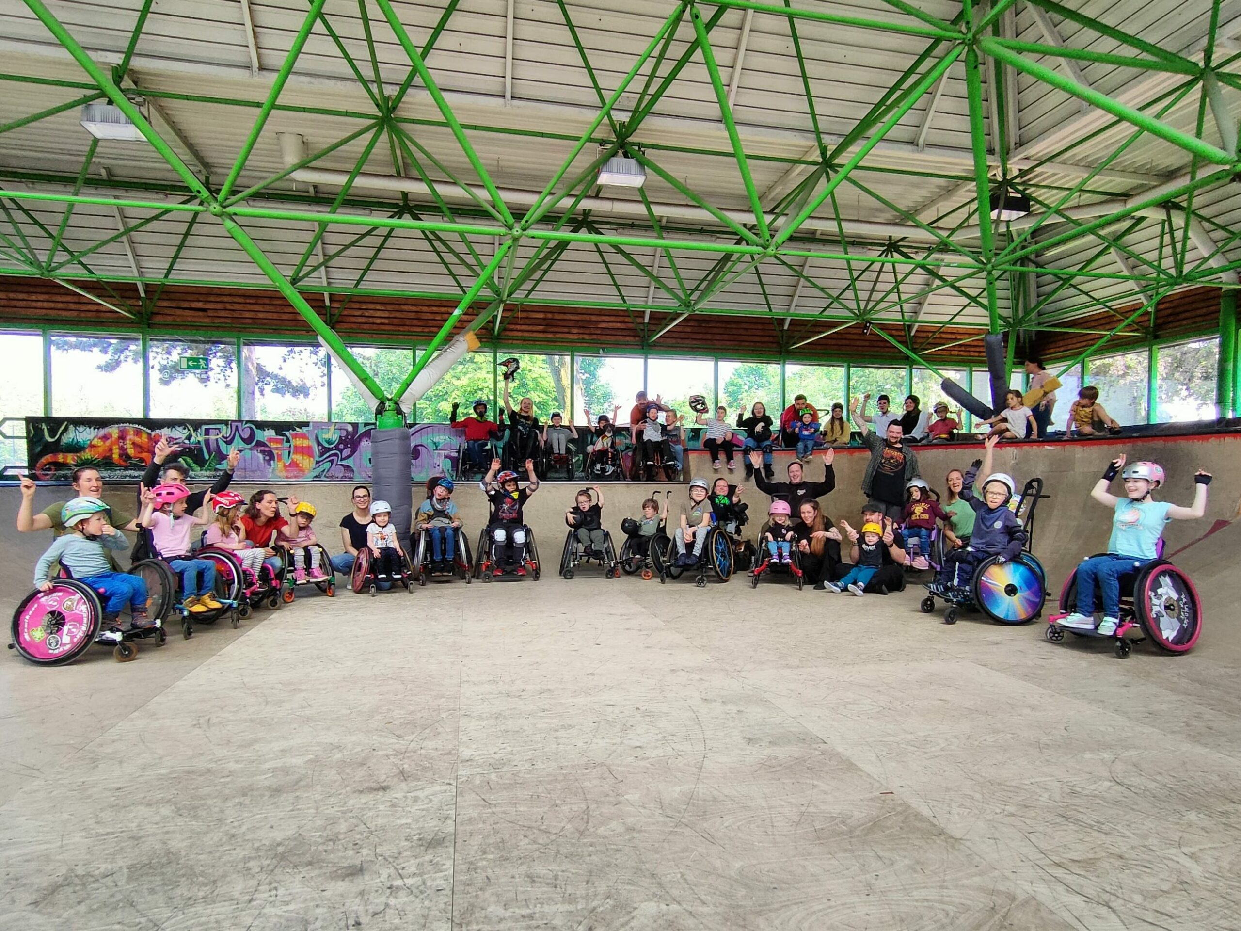 Eine Gruppe von Kindern und Jugendlichem im Rollstuhl in einer Skatehalle. Sie stehen für ein Gruppenfoto auf und vor einer Rampe.