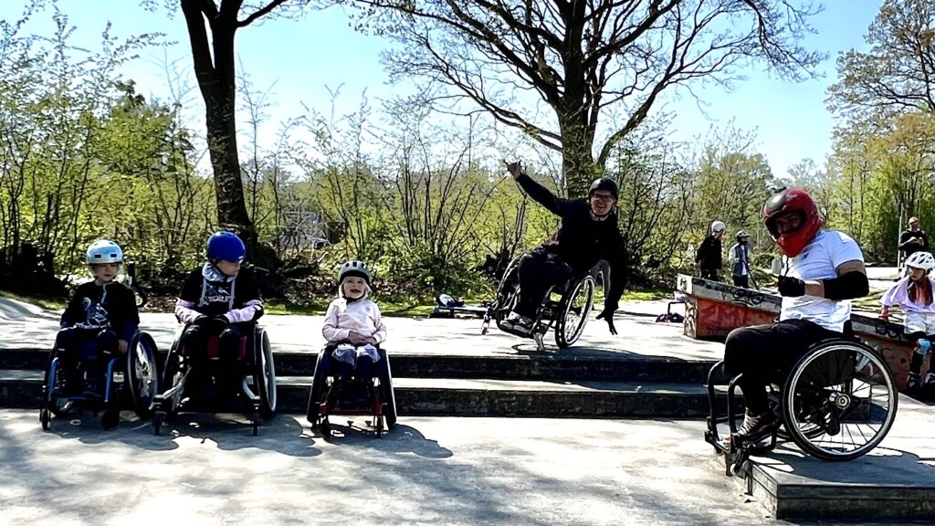 Drei Kinder im Rollstuhl und zwei Erwachsene im Rollstuhl in einem Skatepark. Ein Rollstuhlfahrer balanciert dabei auf einem Rad