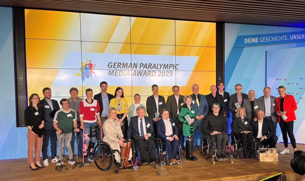 Eine große Gruppe von Menschen mit und ohne Behinderung auf einer Bühne. Auf einem Monitor im Hintergrund steht German Paralympic Media Award.