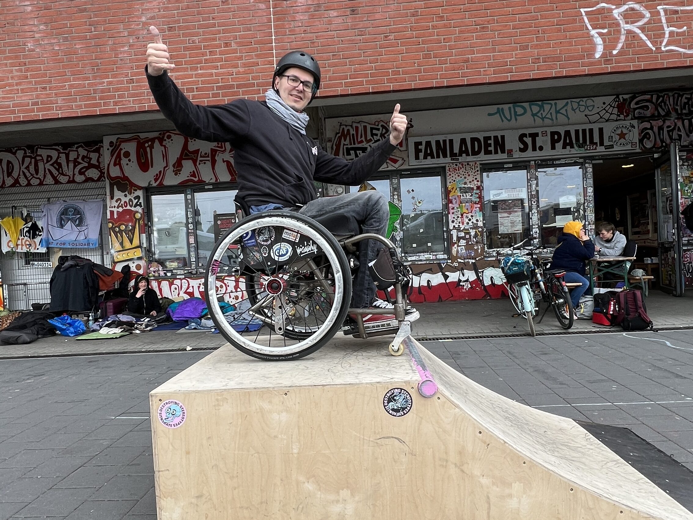 Ein Rollstuhlfahrer steht auf einer mobilen Skaterampe und hält beide Daumen hoch. 
