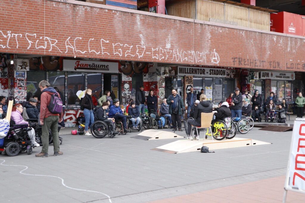 Vor dem Fanladen und den Fanräumen am Millerntorstadion sitzen viele Rollstuhlfahrer und Rollstuhlfahrerinnen, sowie Fußgänger und Fußgängerinnen. Eine Rollstuhlfahrerin sitzt auf einer Holzrampe.