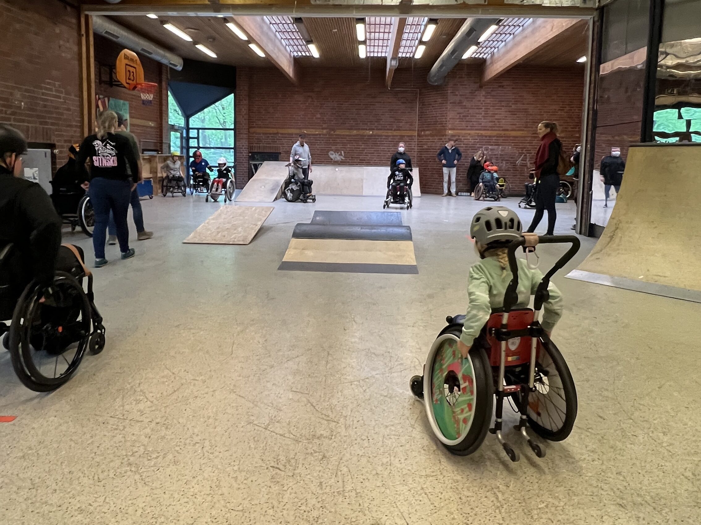 Rollstuhlfahrer und Rollstuhlfahrinnen im Skatepark. Im Vordergrund sieht man ein kleines Mädchen im Rollstuhl, dass Schwung für die Rampen holt. 