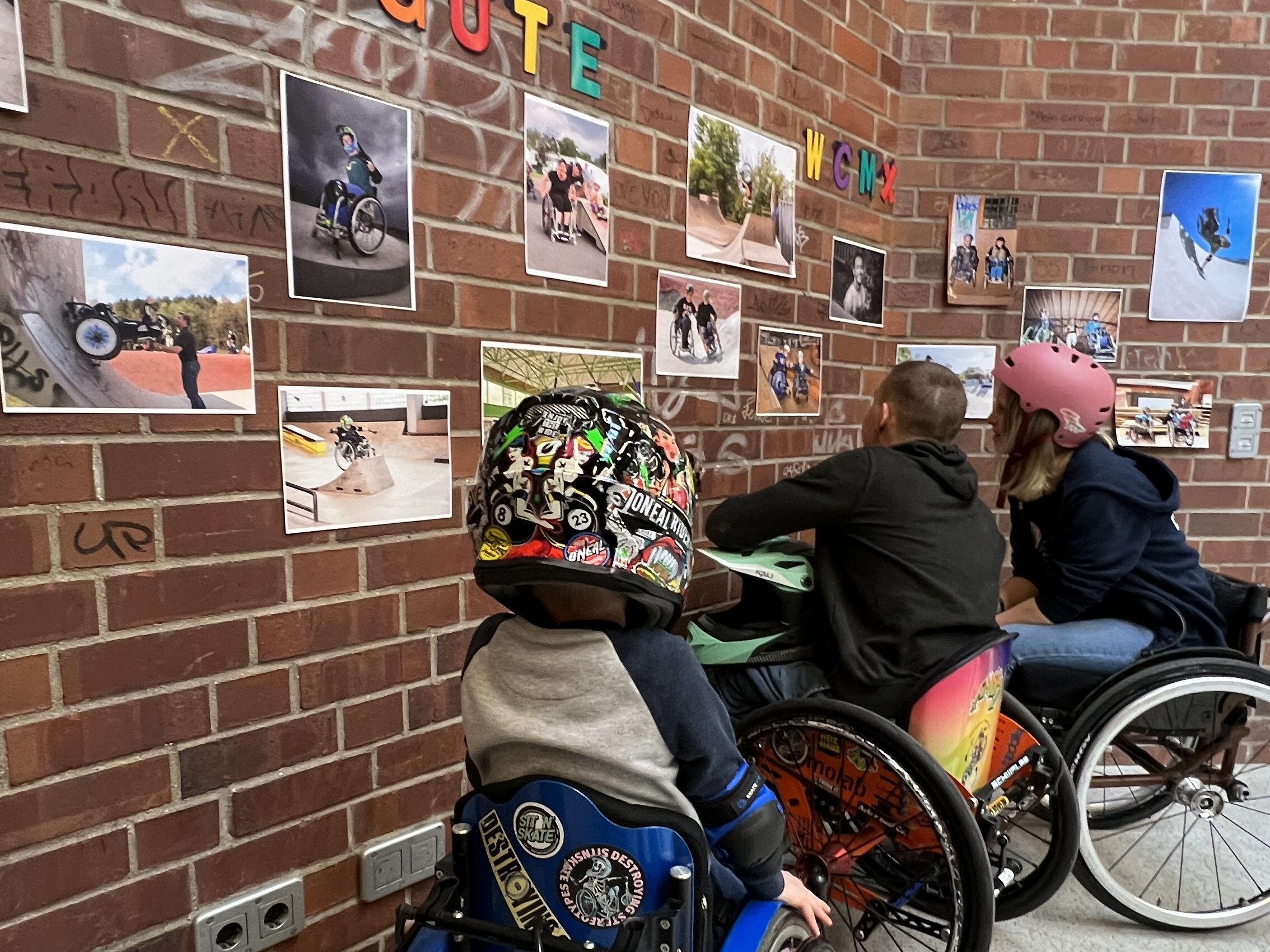 Drei Menschen im Rollstuhl schauen sich Fotos an der Wand an. Auf den Fotos sind Motive von Rollstuhlfahrern und Rolsltuhlfahrerinnen im Skatepark.