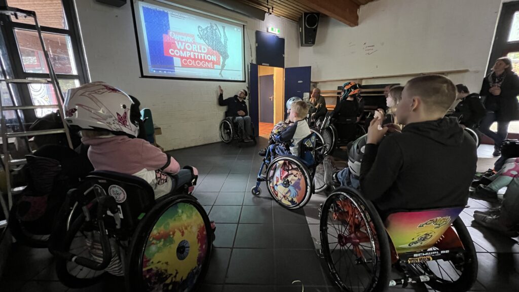 Viele Rollstuhlfahrer und Rollstuhlfahrerinnen schauen auf eine Leinwand. Auf der Leinwand ist das Plakat von der Rollstuhl SKate Weltmeisterschaft zu sehen. 