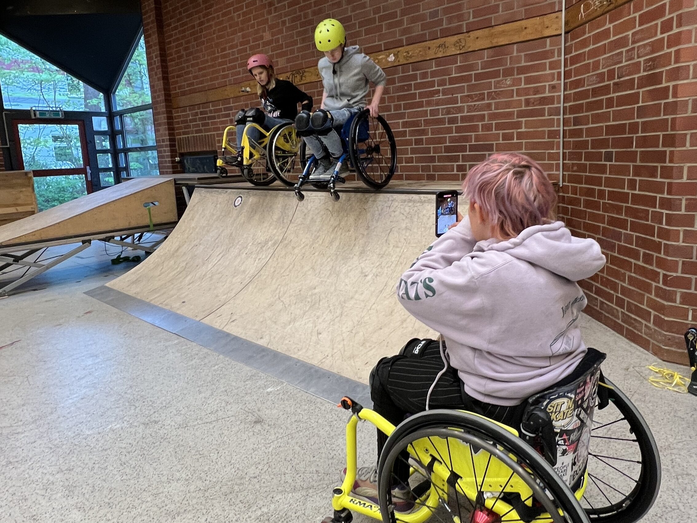 Zwei Rollstuhlfahrerinnen fahren von einer hohen Skaterampe. Im Vordergrund nimmt das eine weitere Rolsltuhlfahrerin auf dem Smartfon auf. 
