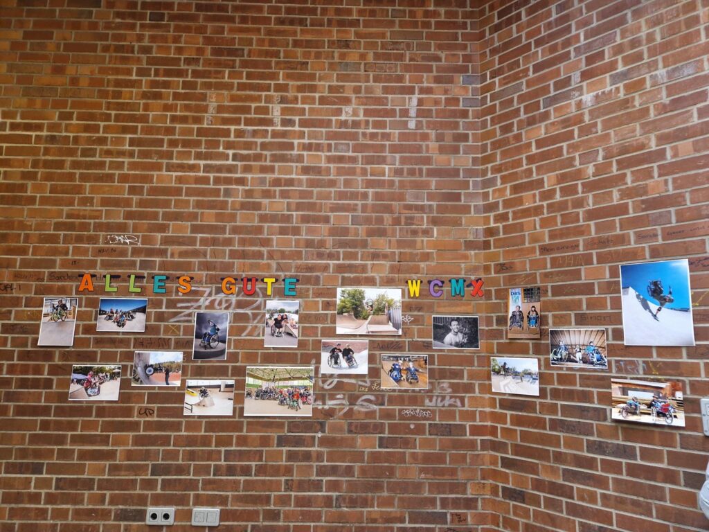 Fotos an einer Wand. Auf den Fotos Motive von Rollstuhlskatern und Rollstuhl Skaterinnen. Darüber steht Alles Gute WCMX. 