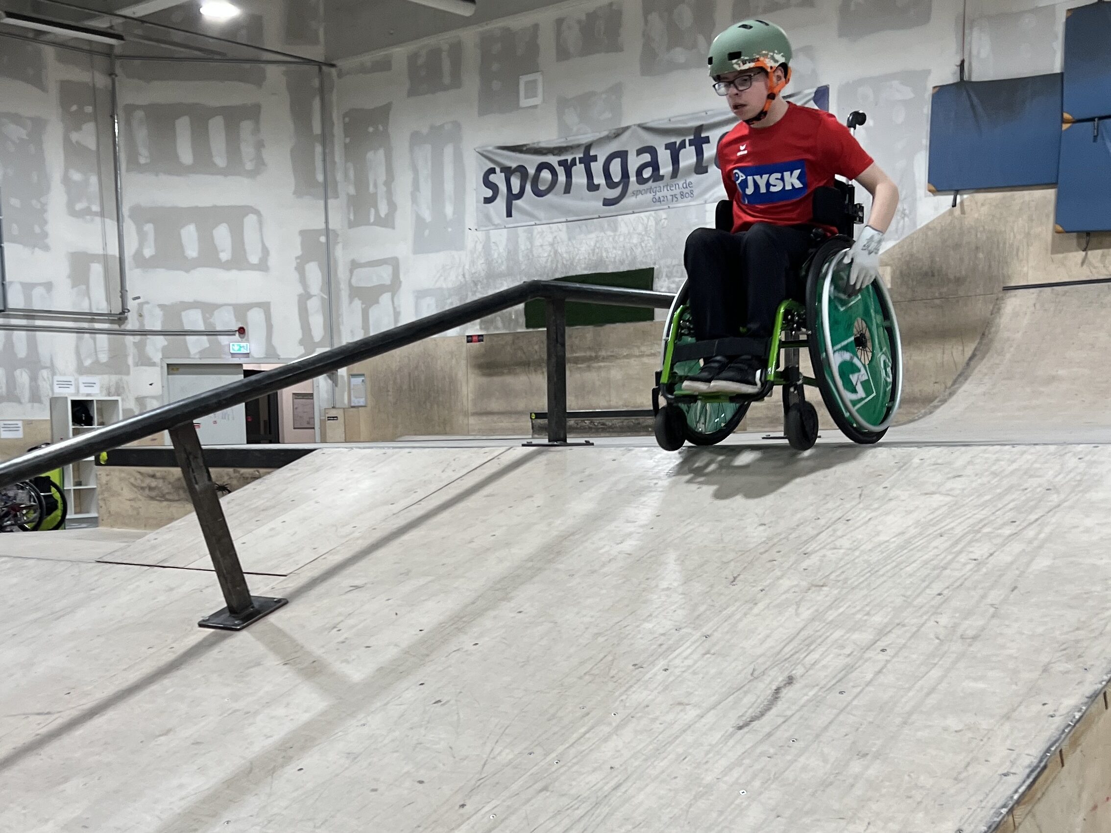 Ein Jugendlicher im Rollstuhl fährt in einer Skatehalle eine Rampe aus Holz runter. 