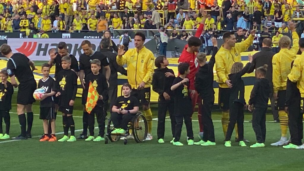 Spieler von Borussia Dortmund stehen auf dem Feld im Stadion. Davor die Einlaufkinder, ein Mädchen davon im Rollstuhl. 