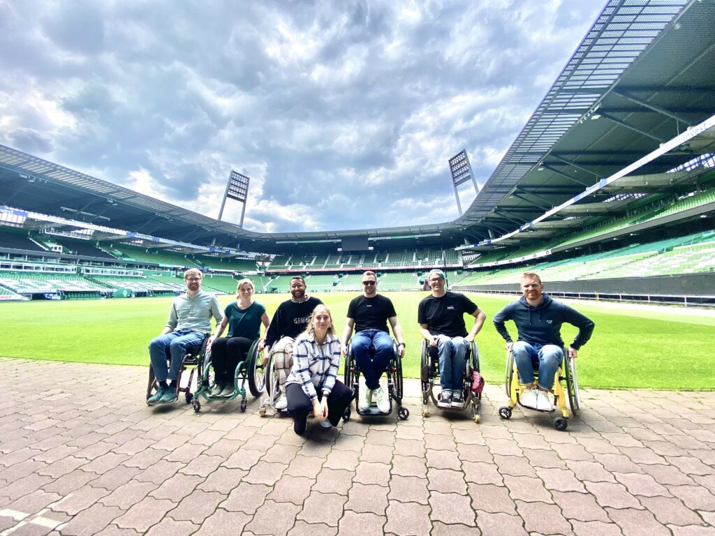 Eine kleine Gruppe von Menschen in Rollstühlen steht in einem leeren Stadion vor dem Rasen.