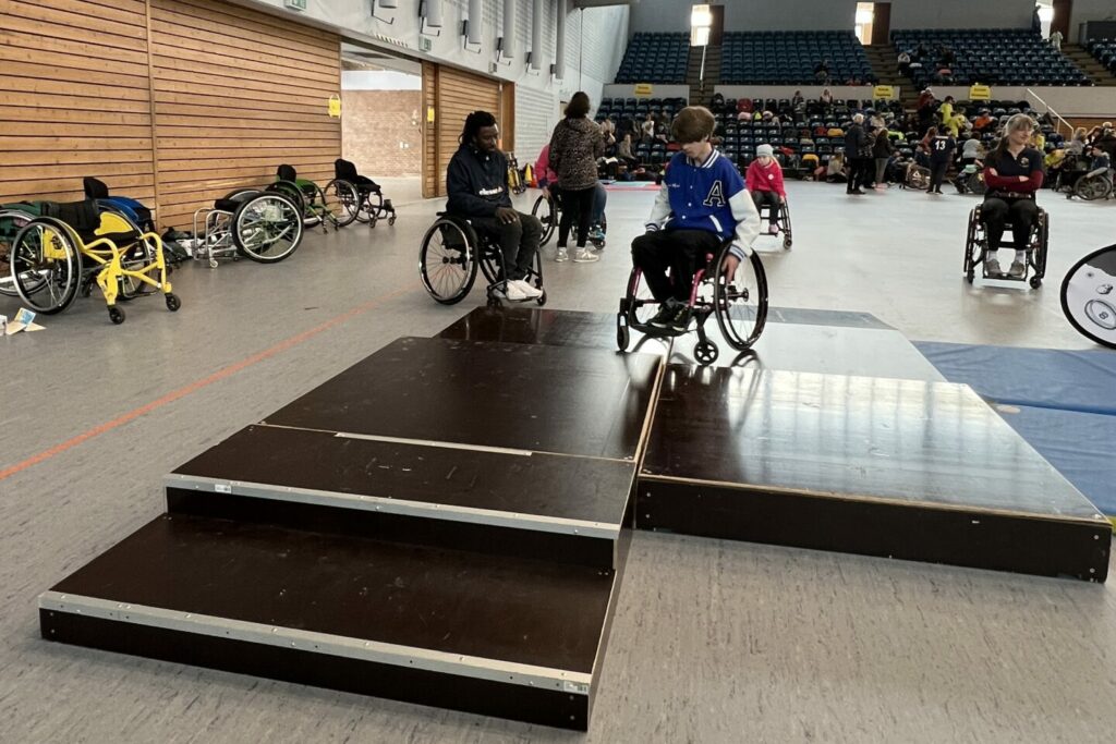 In einer Sporthalle steht ein Parcours aus Holz. Darauf ein Jugendlicher Im Rollstuhl. Weitere Rollstuhlfahrer drum herum